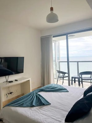 A hotel room with a bed, a TV on a stand, a balcony with chairs, and a view of the sea. - Lujoso Apartamento - Yoo Panamá 21A (Panama City)