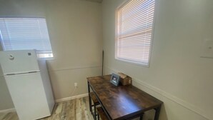 A small kitchen with a wooden table, two chairs, a white refrigerator, and a window with blinds.