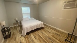 A bedroom with a bed, a nightstand, a small table, a window, and an air conditioning unit.
