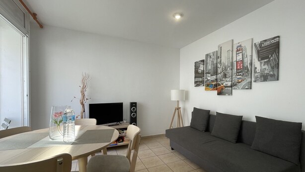 Ein modernes Wohnzimmer mit einem grauen Sofa, einem Esstisch mit Stühlen, einem Fernseher und einem Lautsprecher.
