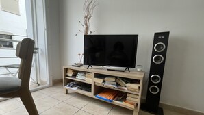 Ein Wohnzimmer mit Fernseher auf einem Holzständer, einer Bücherwand mit Büchern und einem hohen, schwarzen Lautsprecher.