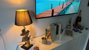 Une pièce avec un téléviseur diffusant un paysage côtier, une lampe, une petite sculpture, une plante dans un pot et une boîte à carreaux.