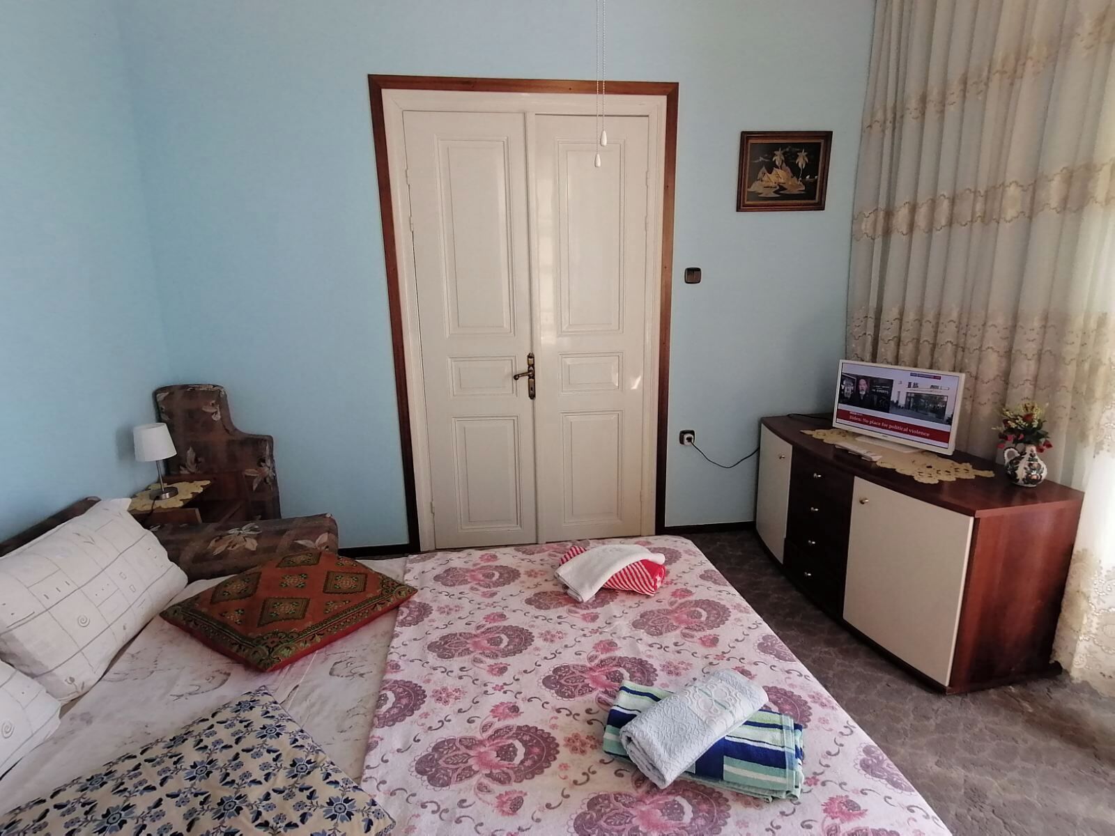 A bedroom with a bed, a wooden headboard, a wardrobe, a desk with a computer, and a curtain.