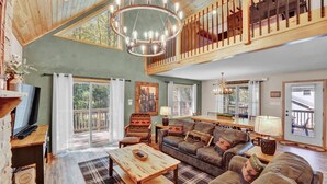 A spacious living room with a high ceiling, a wooden ceiling, a large chandelier, a fireplace, a television, a dining table, and a sofa with decorative pillows. - Emerald Bliss, MiniGolf Bowling Playset Beach Horseshoe Cornhole (Towamensing Trails)