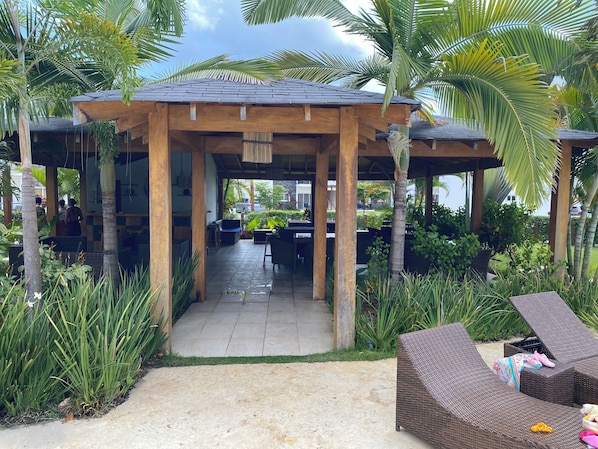 A covered outdoor seating area with tables and chairs, surrounded by lush greenery and palm trees.