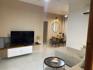 A modern living room with a flat-screen TV, a dining area with a table and chairs, a round mirror, and a small round table with a book.