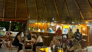 Un restaurante al aire libre con mesas y sillas de madera, techo de paja y zona de bar con gente cenando.