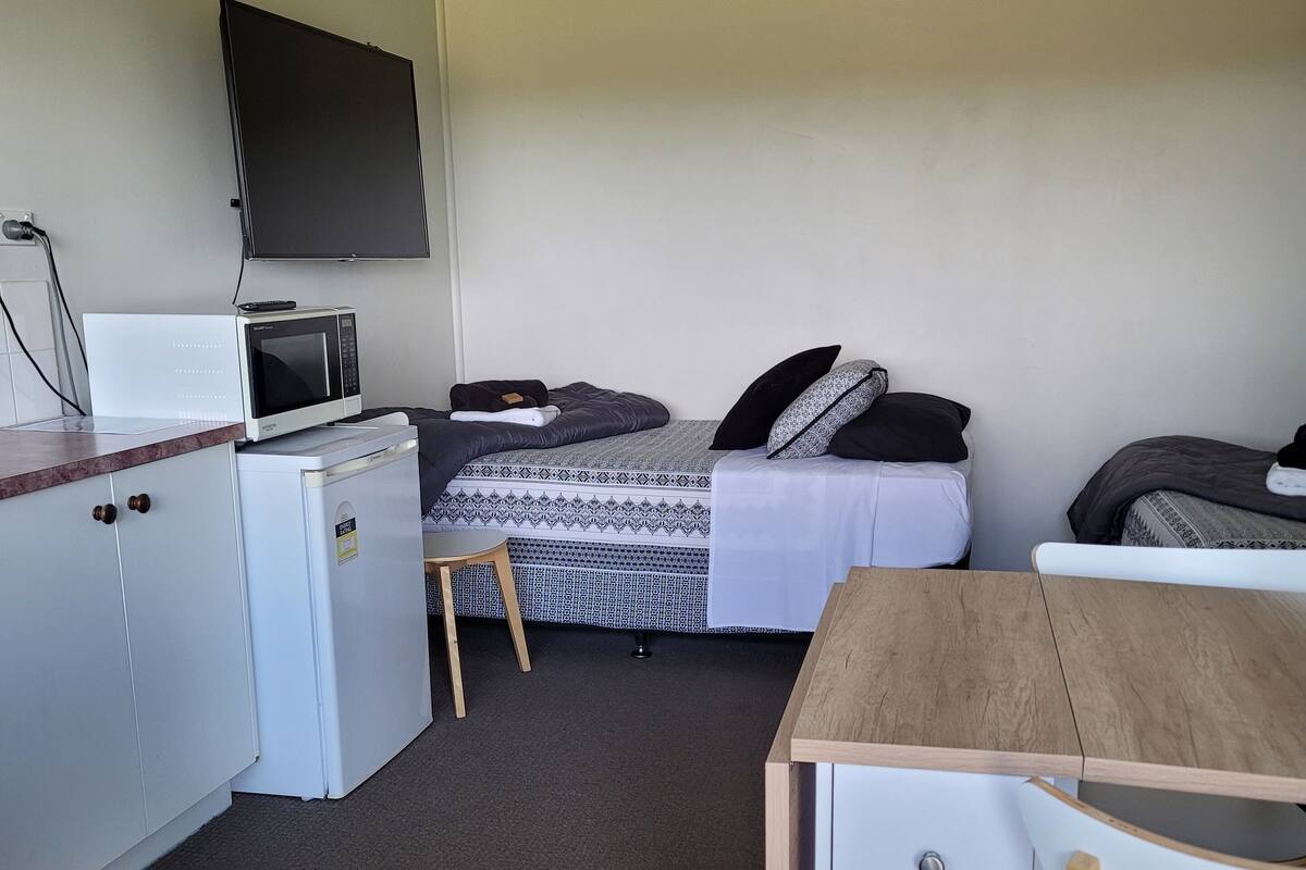 A compact room with a kitchenette, a bed with patterned bedding, a TV mounted on the wall, and a small wooden table.