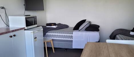 A compact room with a kitchenette, a bed with patterned bedding, a TV mounted on the wall, and a small wooden table.