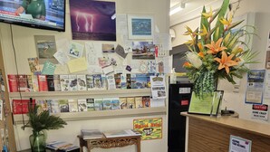 A reception area with a wooden desk, a computer monitor, a bulletin board with various notices, a small table, and a potted plant. - Jabiru Motel (Nambucca Heads)