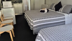 A compact hotel room with a kitchenette, two beds with patterned bedding, a desk with a chair, and a wardrobe. - Jabiru Motel (Nambucca Heads)