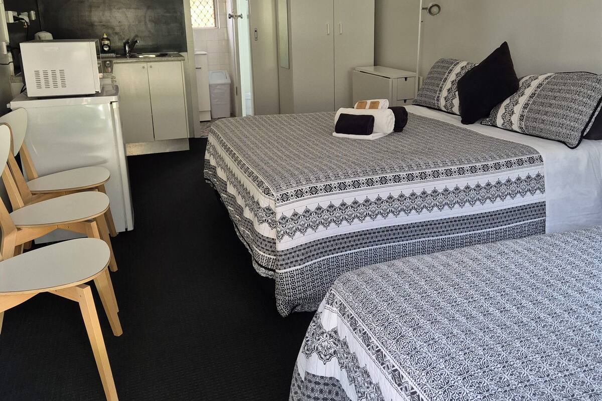A compact hotel room with a kitchenette, two beds with patterned bedding, a desk with a chair, and a wardrobe.