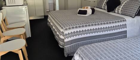Habitación de hotel compacta con kitchenette, dos camas con ropa de cama estampada, un escritorio con silla y un armario.