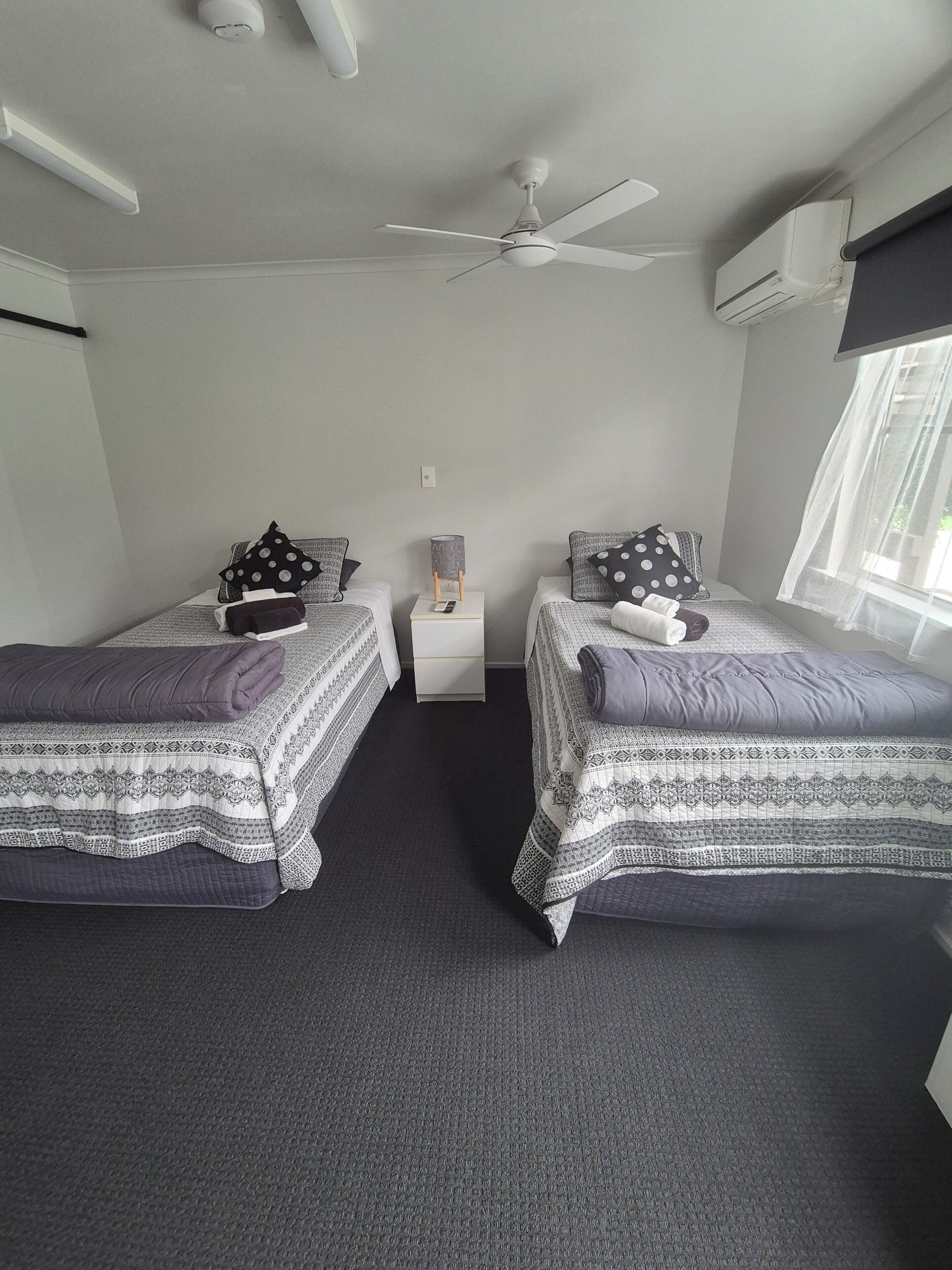 A room with two beds, a ceiling fan, a small table with a lamp, and a window with sheer curtains.