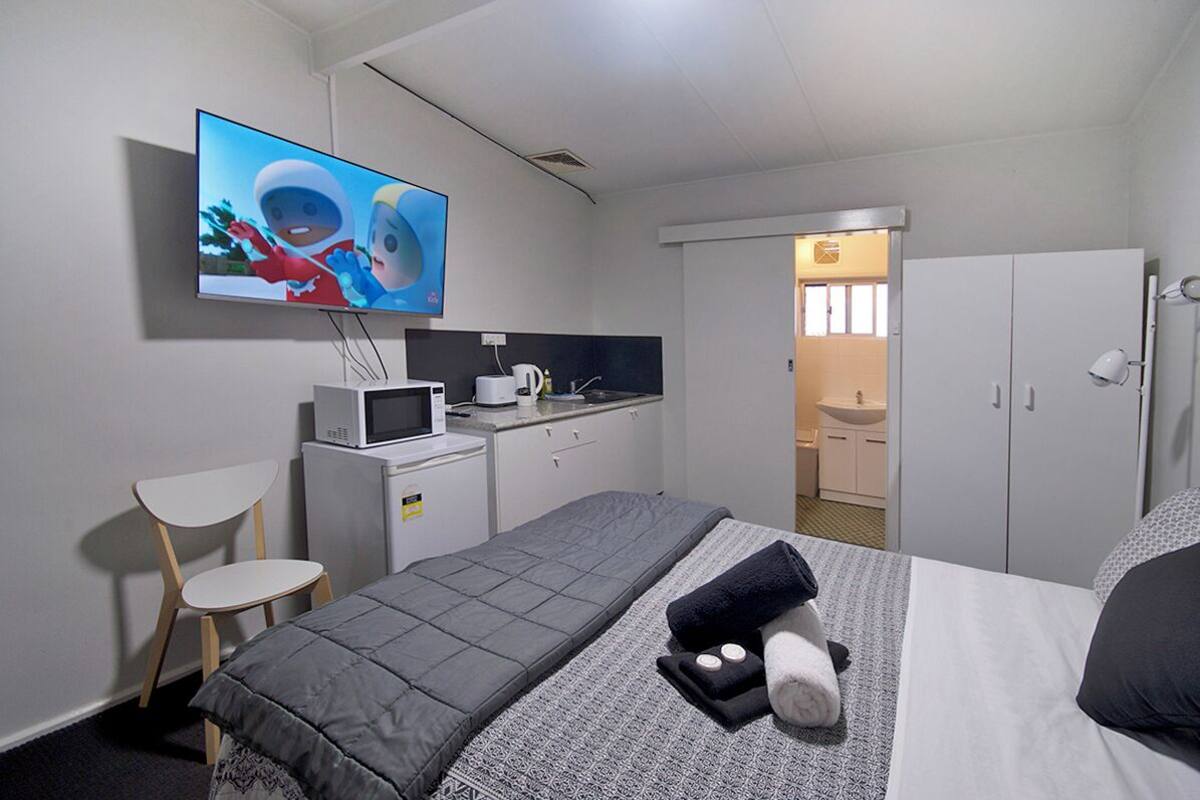 A hotel room with a bed, a TV mounted on the wall, a small kitchenette, a desk, and a chair.