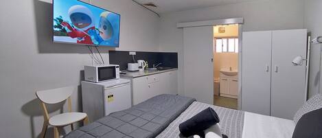 Habitación de hotel con cama, televisor montado en la pared, kitchenette pequeña, escritorio y una silla.