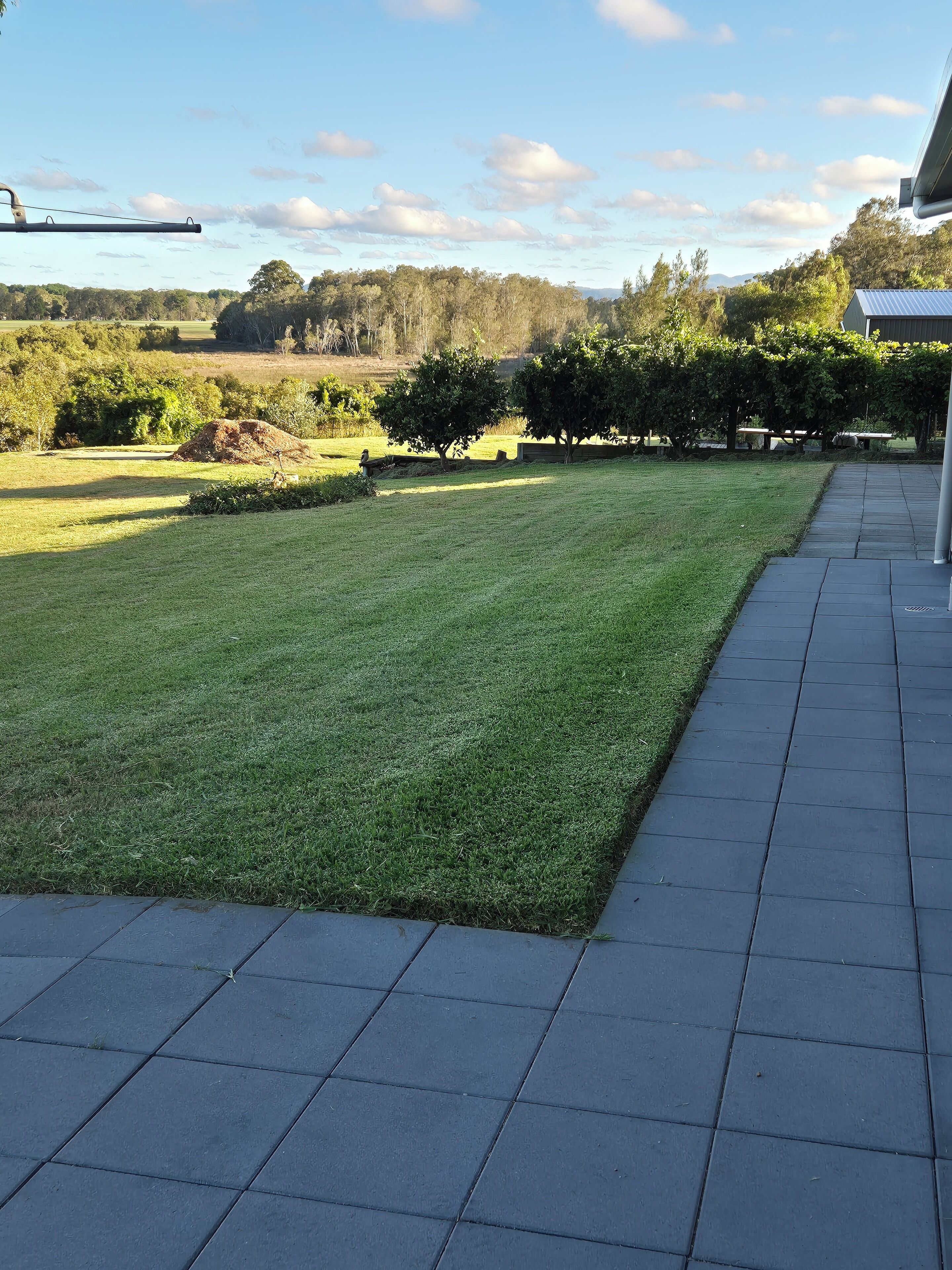 A well-maintained lawn with a paved walkway and a view of trees and hills in the distance.