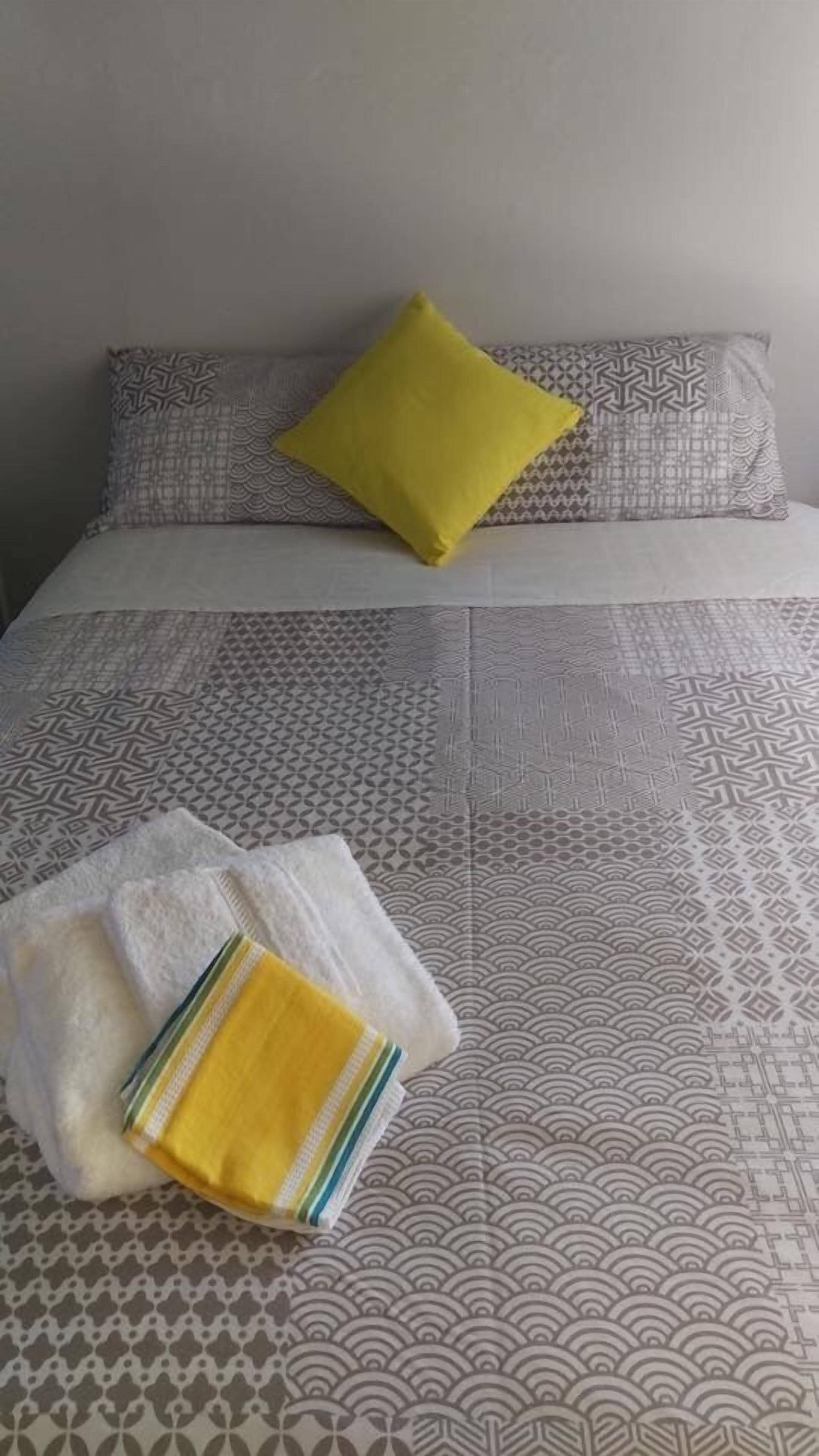 A bed with patterned bedding, a yellow pillow, and a folded towel with a striped towel on it.