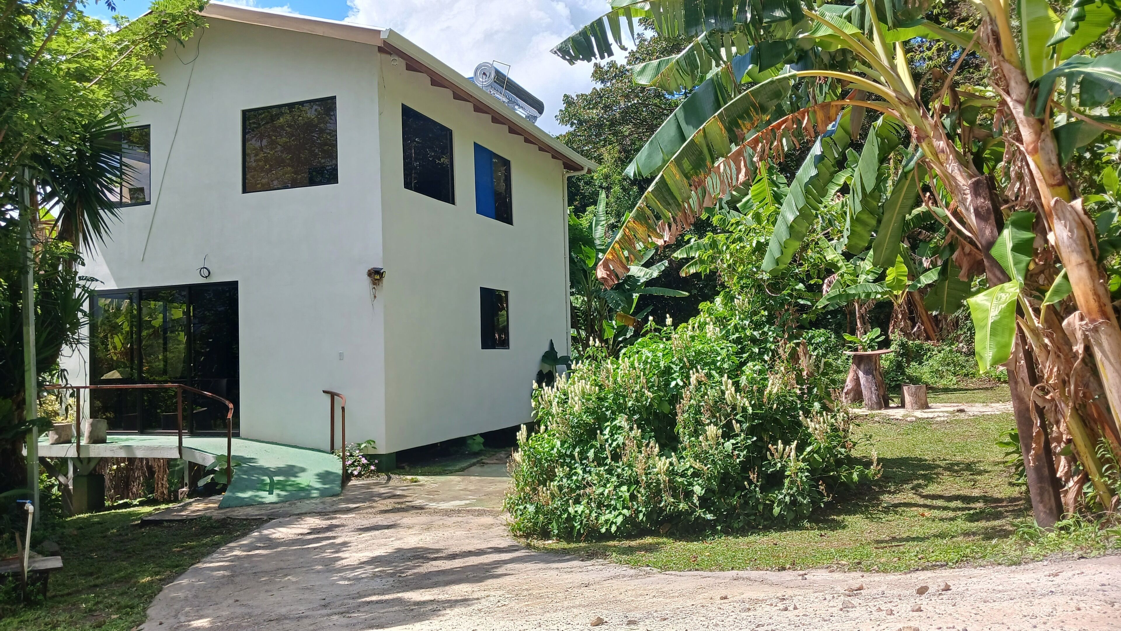 A white two-story house with a small porch, surrounded by lush greenery and banana trees.