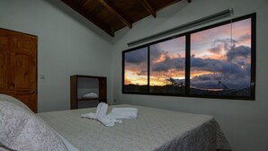Un dormitorio con un amplio ventanal que muestra una vista al atardecer, una cama con una manta gris y una puerta de madera.