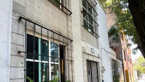 Un edificio de dos plantas con ventanas amplias y una puerta, rodeado de árboles y una acera.