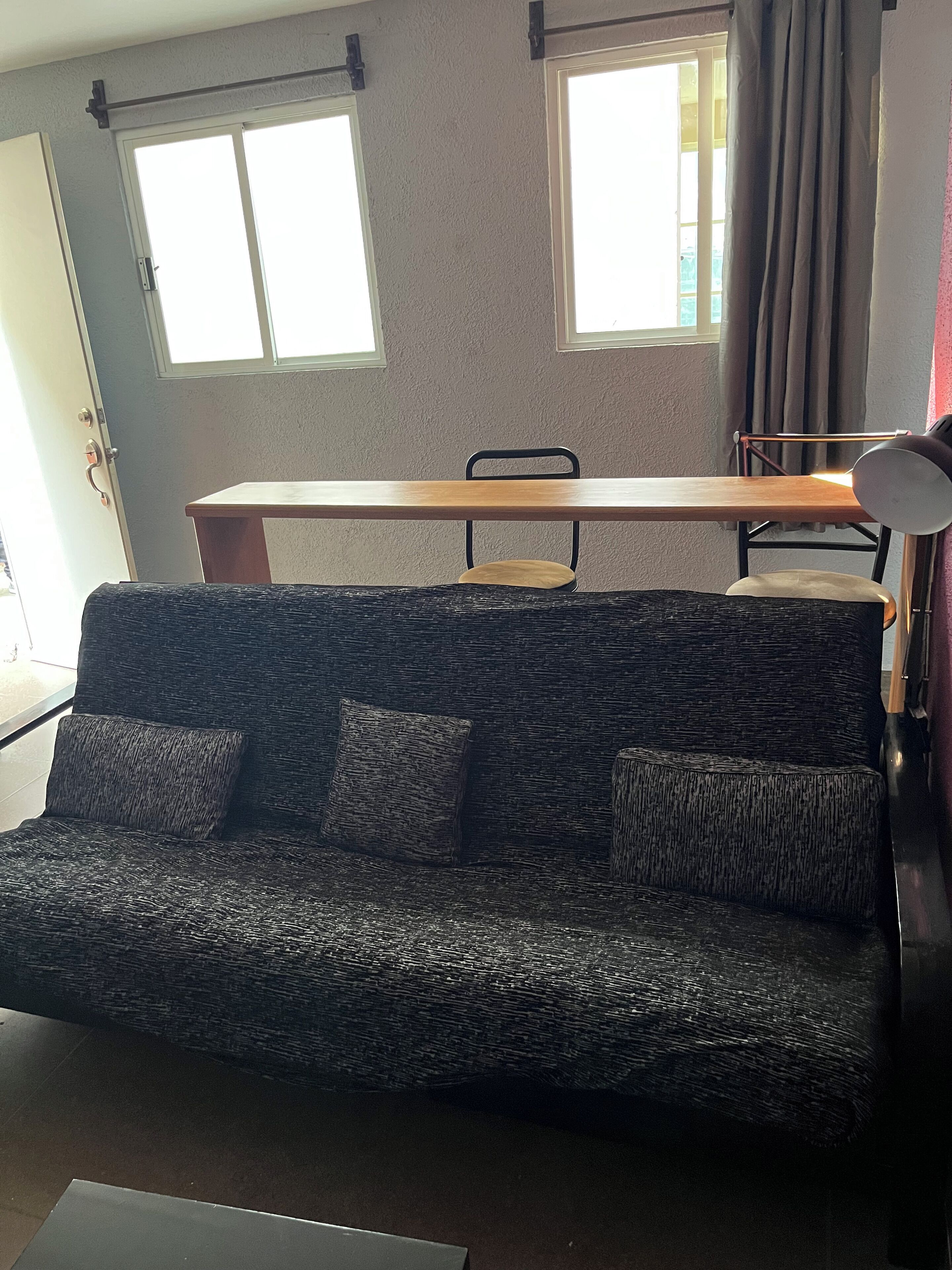 Una sala de estar con un sofá de color oscuro y estampado, una mesa de comedor de madera y una silla. Hay dos ventanas con cortinas, una puerta y una lámpara sobre un pedestal.