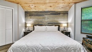 Una camera da letto con soffitto in legno, un letto grande con biancheria bianca, due comodini con lampade e una finestra con persiane, più una parete decorata.