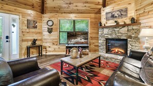 Interno di una baita in legno con camino in pietra, divani in pelle, un tavolo di legno e un'ampia finestra con persiane.
