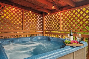 A hot tub with a wooden deck and lattice panels, a bottle of wine, and a tray with grapes and glasses.