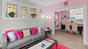 A living room with a grey sofa, a coffee table, and a dining area with a pink wall and heart-pattern wallpaper.