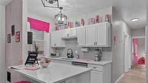 A modern kitchen with white cabinets, a central island with pink stools, a 'Welcome to Barbie Dreamhouse' sign, and a window with blinds.