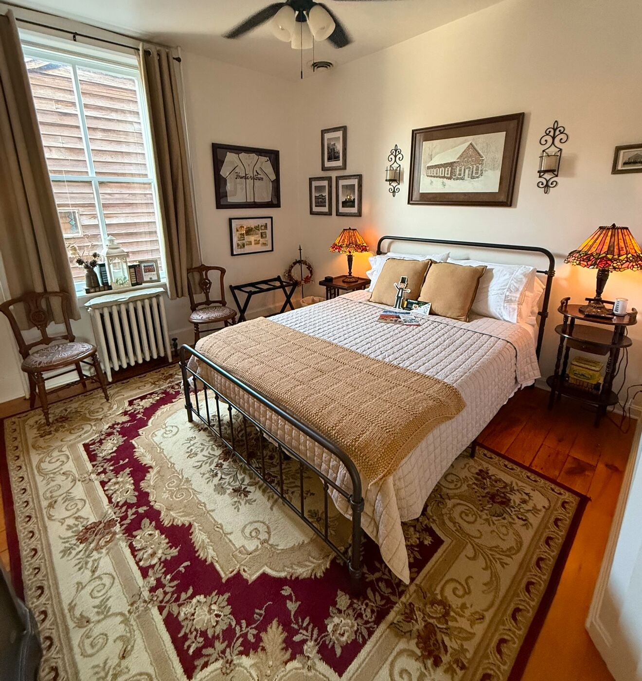 A bedroom with a bed, a radiator, a window with curtains, a chair, a lamp, and wall decorations.
