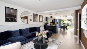 A modern living room with a dark blue sofa, a round coffee table with a vase of flowers, and a large mirror on the wall. - Stunning 4BR 3BA in the ♥️ of London ❄️ Air Con. 🌿 Private Garden | Sleeps 10 (Greater London)