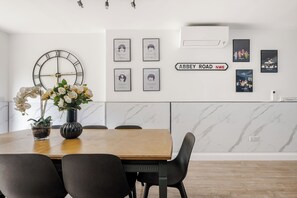 A dining area with a wooden table, black chairs, a vase with flowers, a wall clock, framed pictures, and a sign reading 'ABBEY ROAD NW8'.