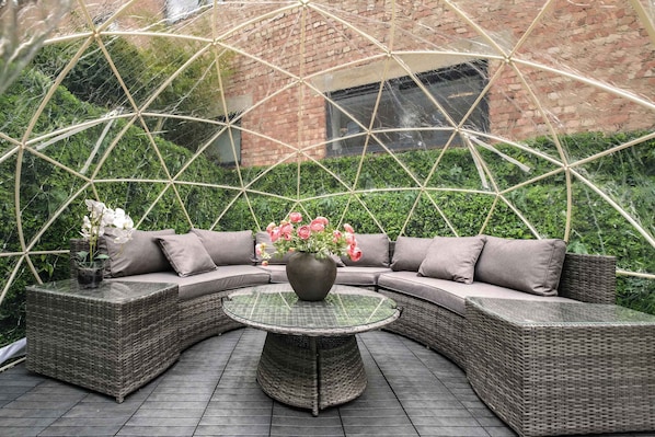 A covered outdoor seating area with a round table, cushioned sofa, and a potted plant.