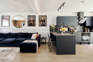 A modern kitchen with dark cabinetry, a central island, and a dining area with a dark blue sofa. - Stunning 4BR 3BA in the ♥️ of London ❄️ Air Con. 🌿 Private Garden | Sleeps 10 (Greater London)