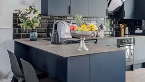 A modern kitchen with dark cabinetry, a central island with a fruit bowl, and a vase of flowers. - Stunning 4BR 3BA in the ♥️ of London ❄️ Air Con. 🌿 Private Garden | Sleeps 10 (Greater London)