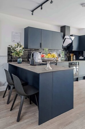 A modern kitchen with dark cabinetry, a central island with a fruit bowl, and a vase of flowers. - Stunning 4BR 3BA in the ♥️ of London ❄️ Air Con. 🌿 Private Garden | Sleeps 10 (Greater London)