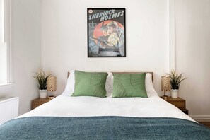 Quarto com cama, almofadas verdes, pôster emoldurado na parede e duas mesas de cabeceira com luminárias e plantas.