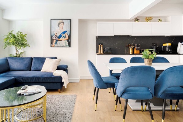 A modern living room with a blue sofa, a glass coffee table, and a dining area with blue chairs and a white table.