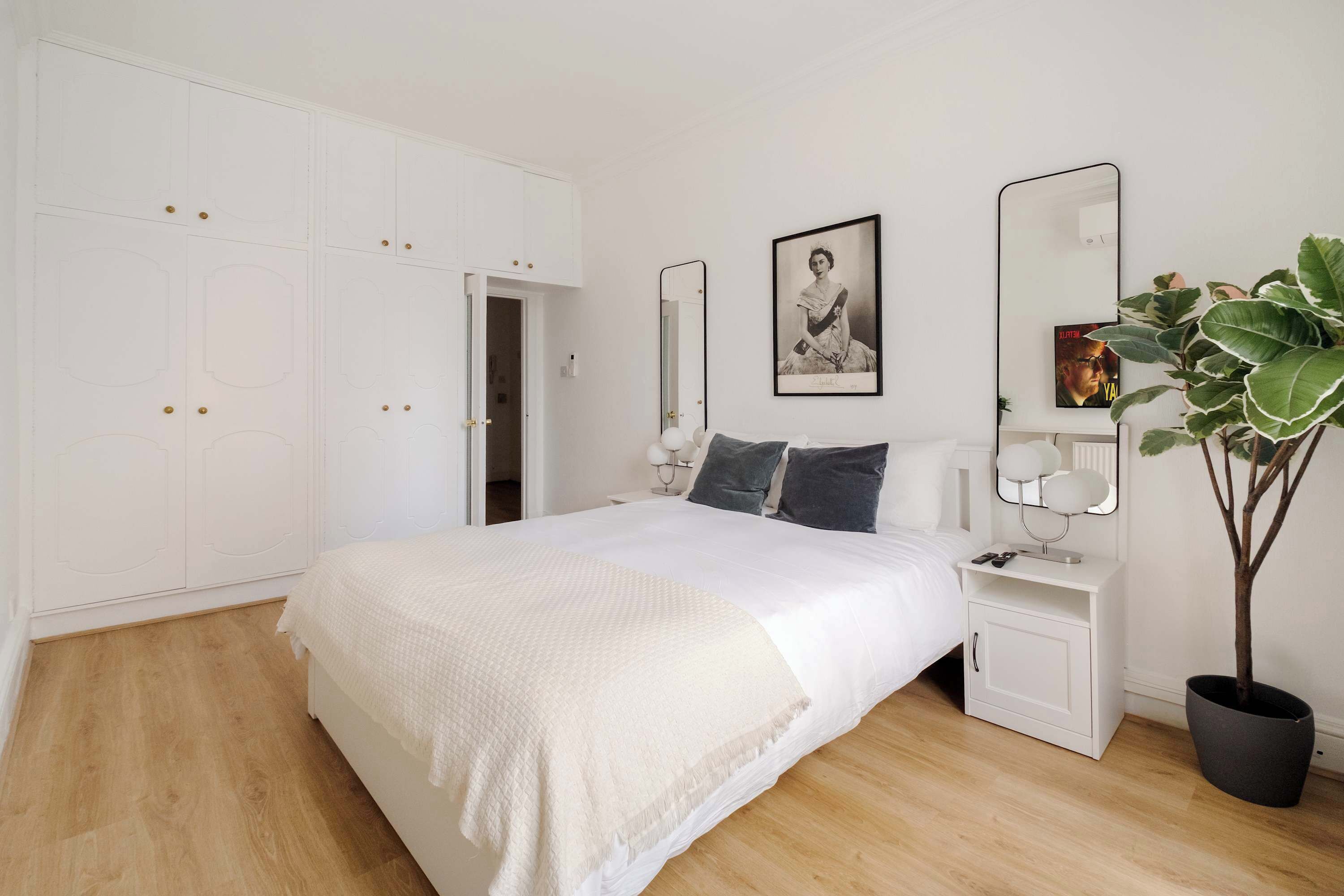 A bedroom with a large bed, white wardrobe, bedside tables with lamps, and a potted plant.