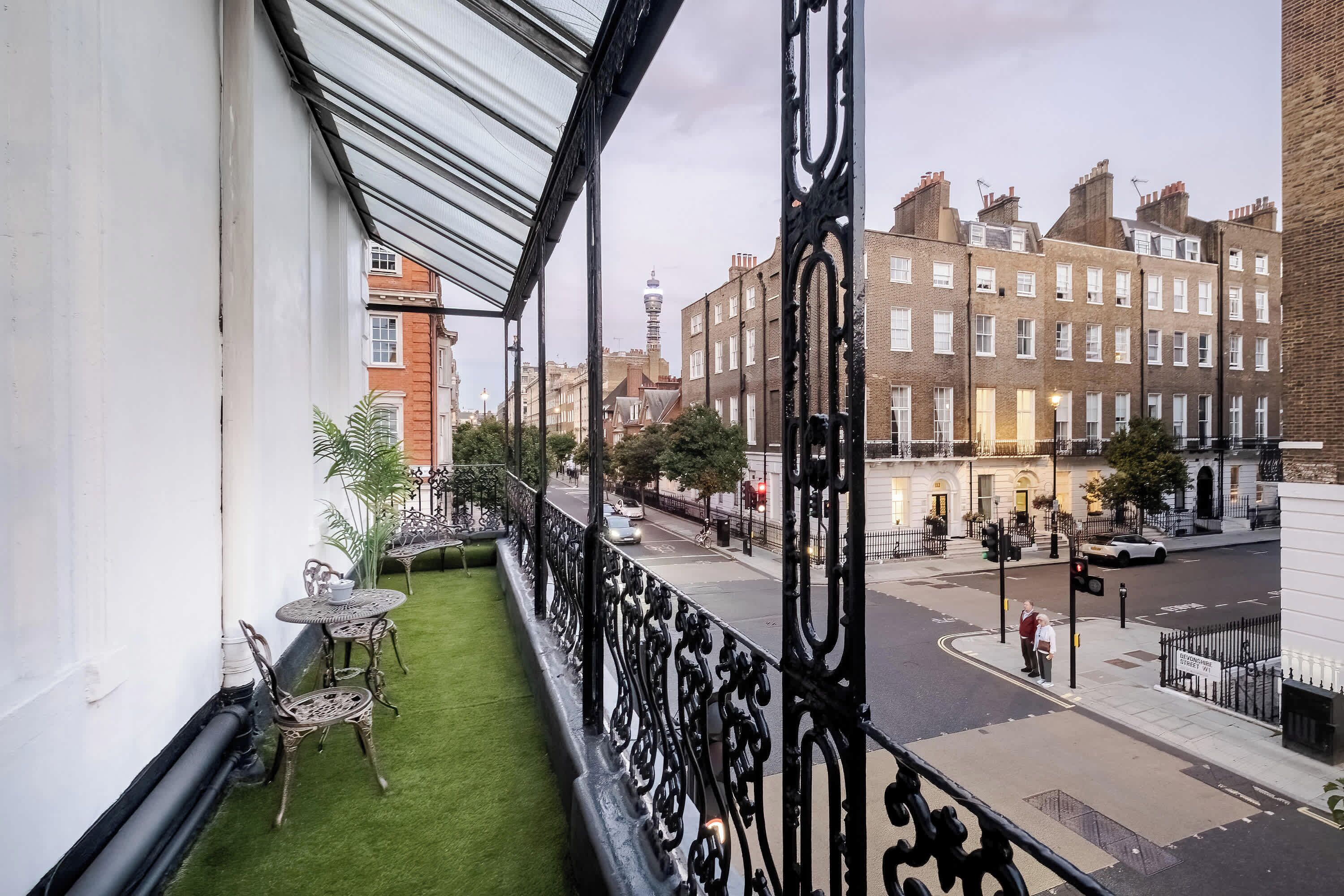 A balcony with a view of a city street, a small garden area with chairs, and a white building in the background.