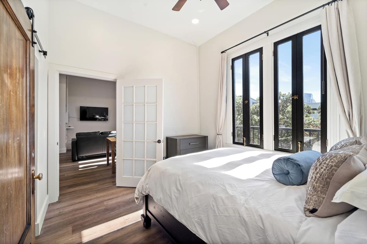 A bedroom with a large bed, a TV in the adjacent room, and a balcony with a view.