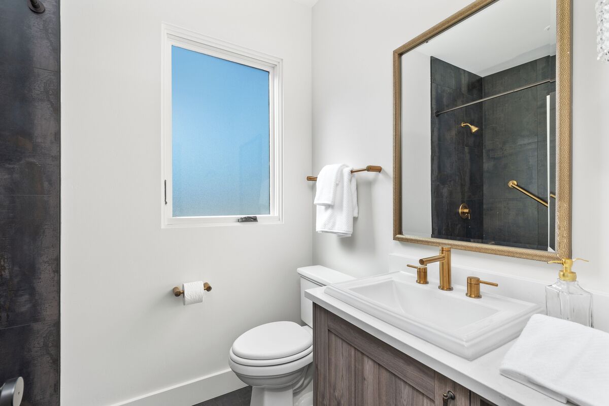 Et badeværelse med et hvidt toilet, en hvid vask med guldfarvede armaturer, et stort spejl og et vindue med udsigt til himlen.