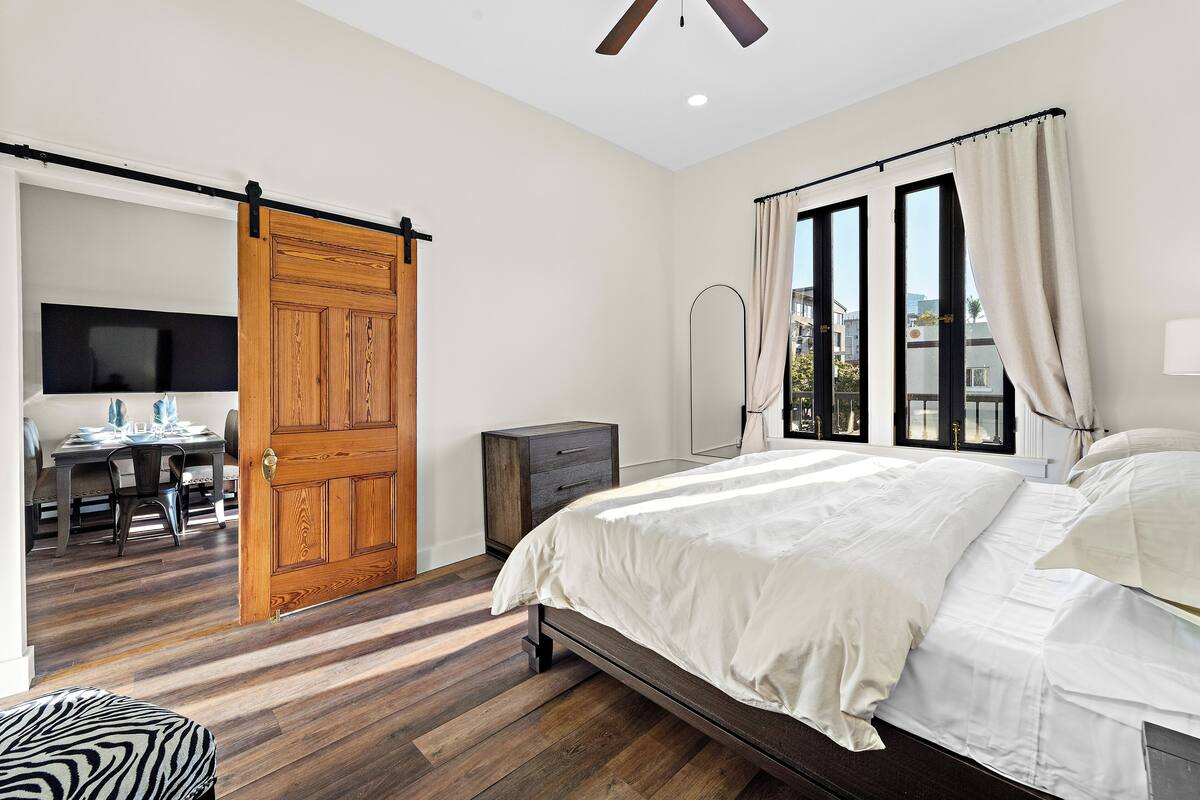 A bedroom with a bed, a wooden door, a desk with a chair, and a window with curtains.
