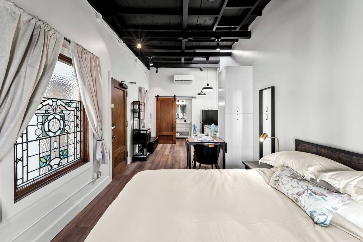A bedroom with a large bed, a desk, and a chair. There is a window with curtains and a door leading to another room.