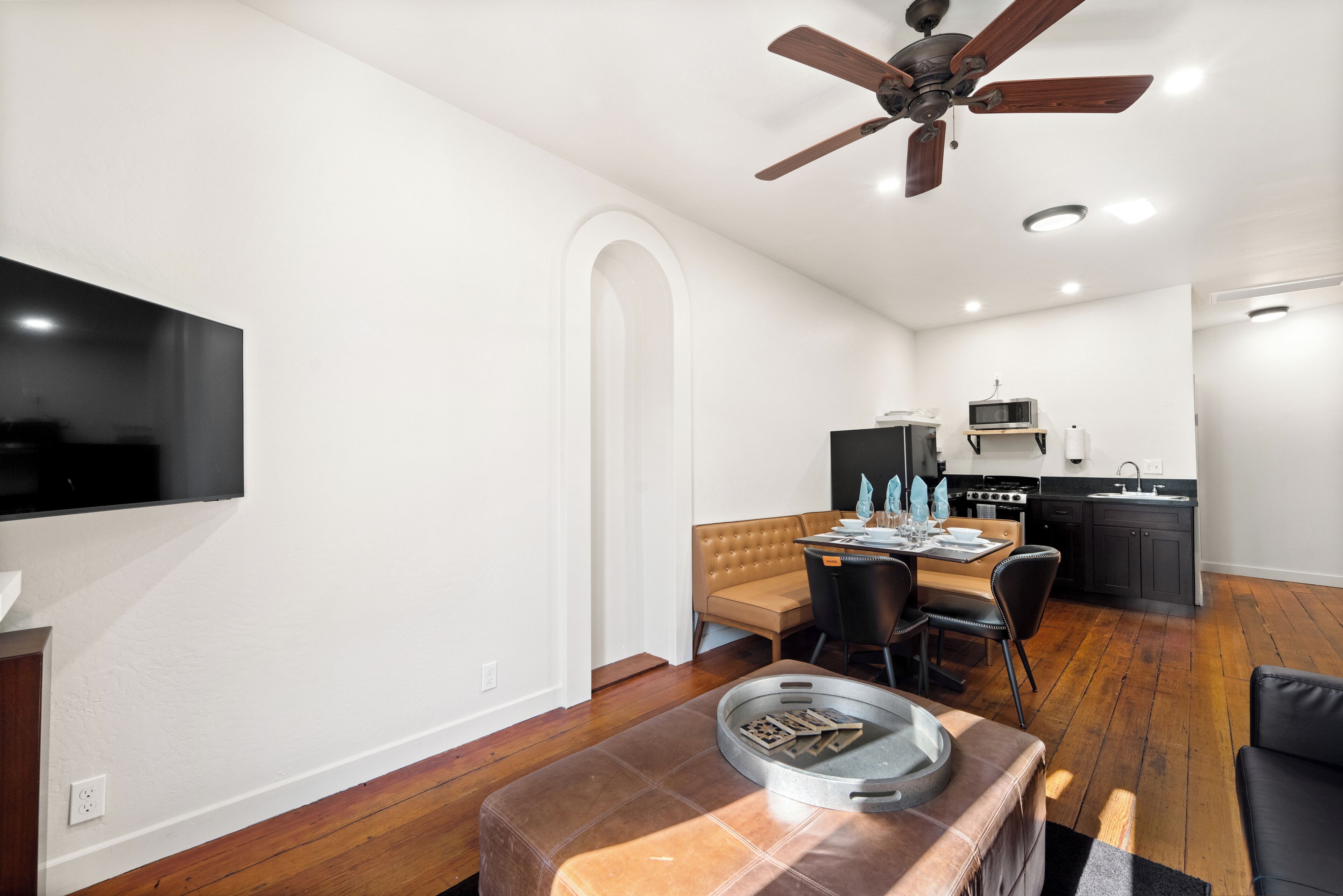 A modern living room with a flat-screen TV, a dining area with a table and chairs, a kitchen with a stove and microwave, and a ceiling fan.
