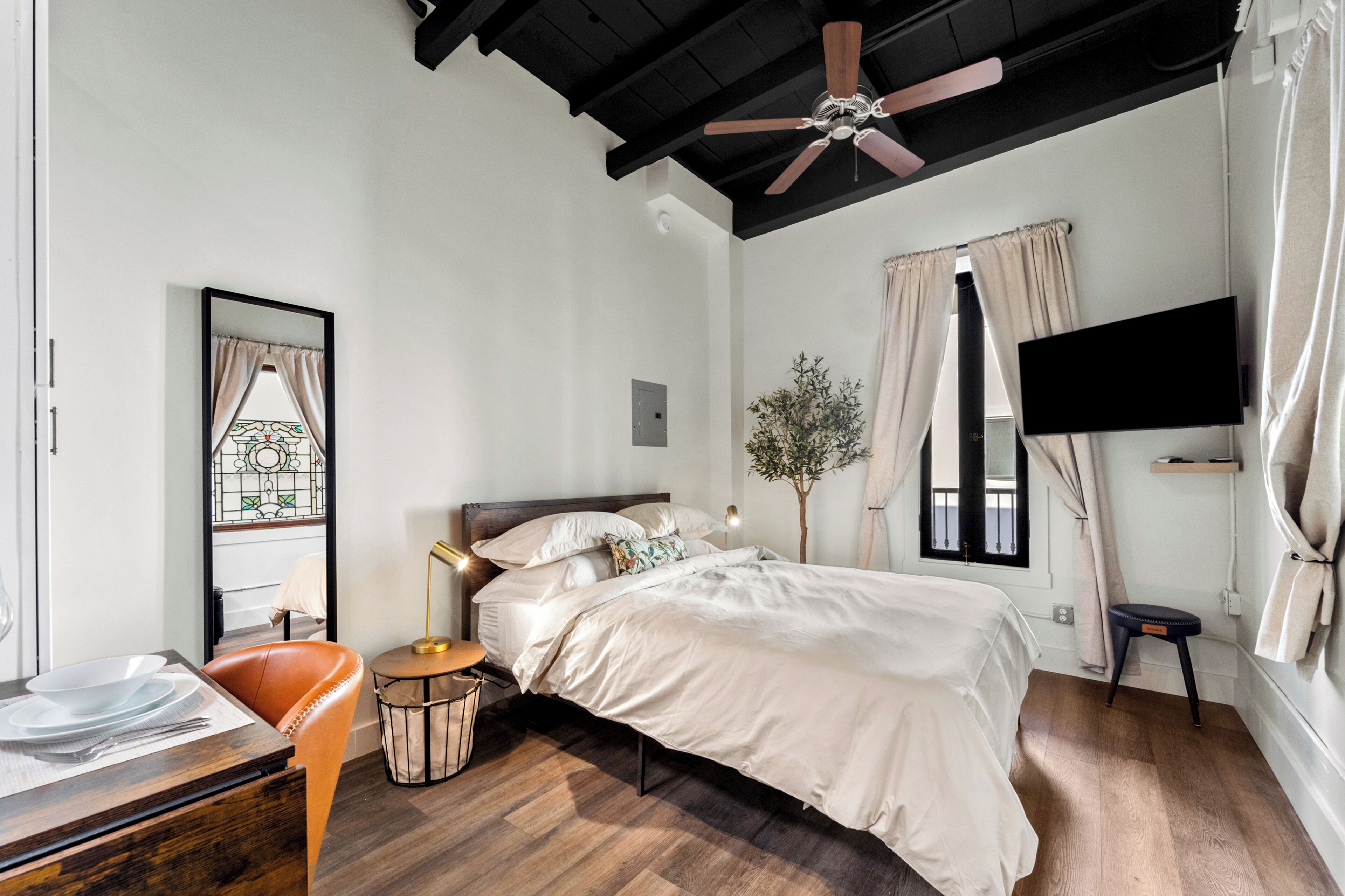 A bedroom with a bed, a desk with a chair, a ceiling fan, a TV mounted on the wall, and a window with curtains.