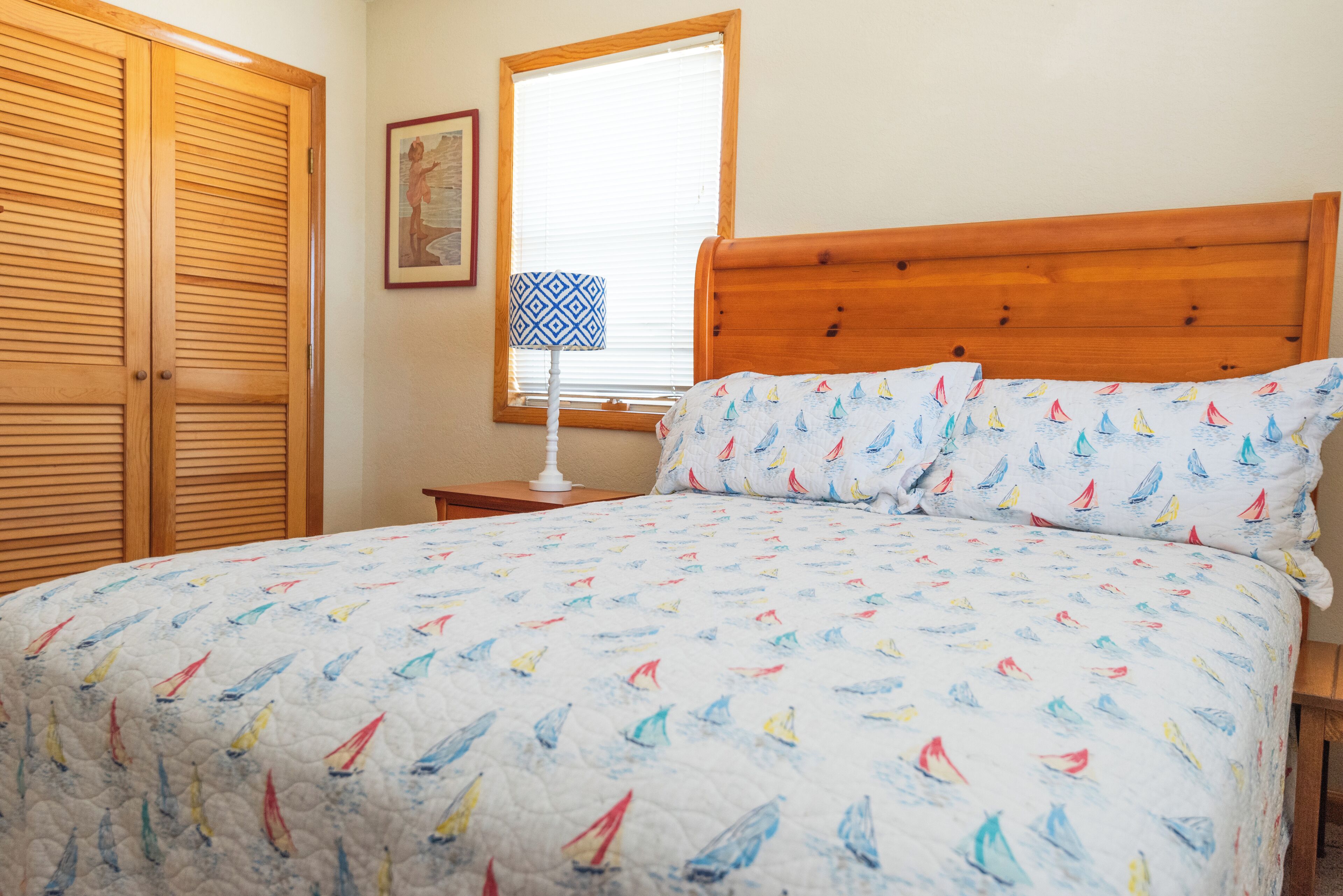 Un dormitorio con cabecera de madera, cama con edredón estampado de velero, mesita de noche con lámpara y ventana con persianas.