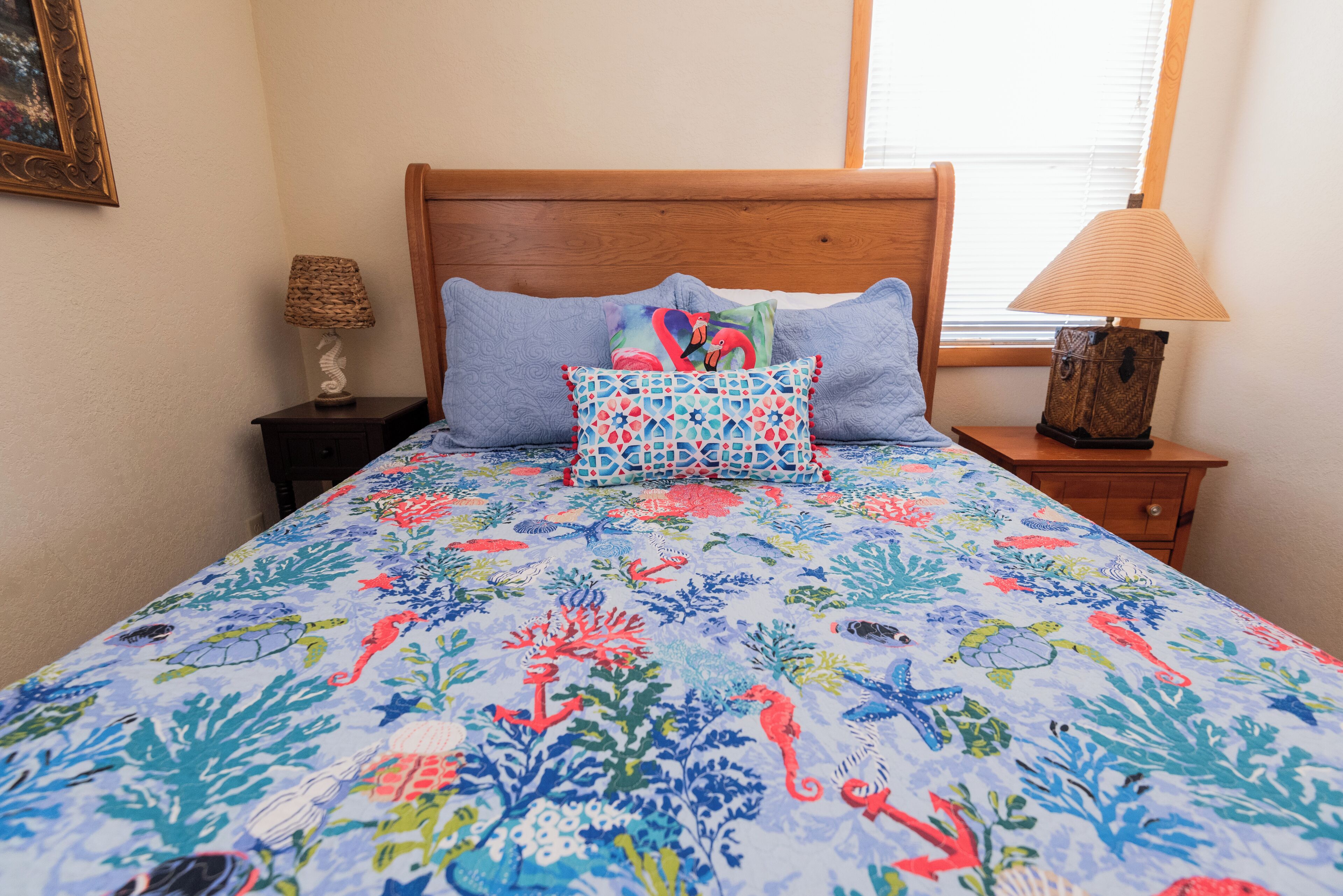 A bed with a colorful floral bedspread, two pillows, a wooden headboard, a nightstand with a lamp, and a window with blinds.